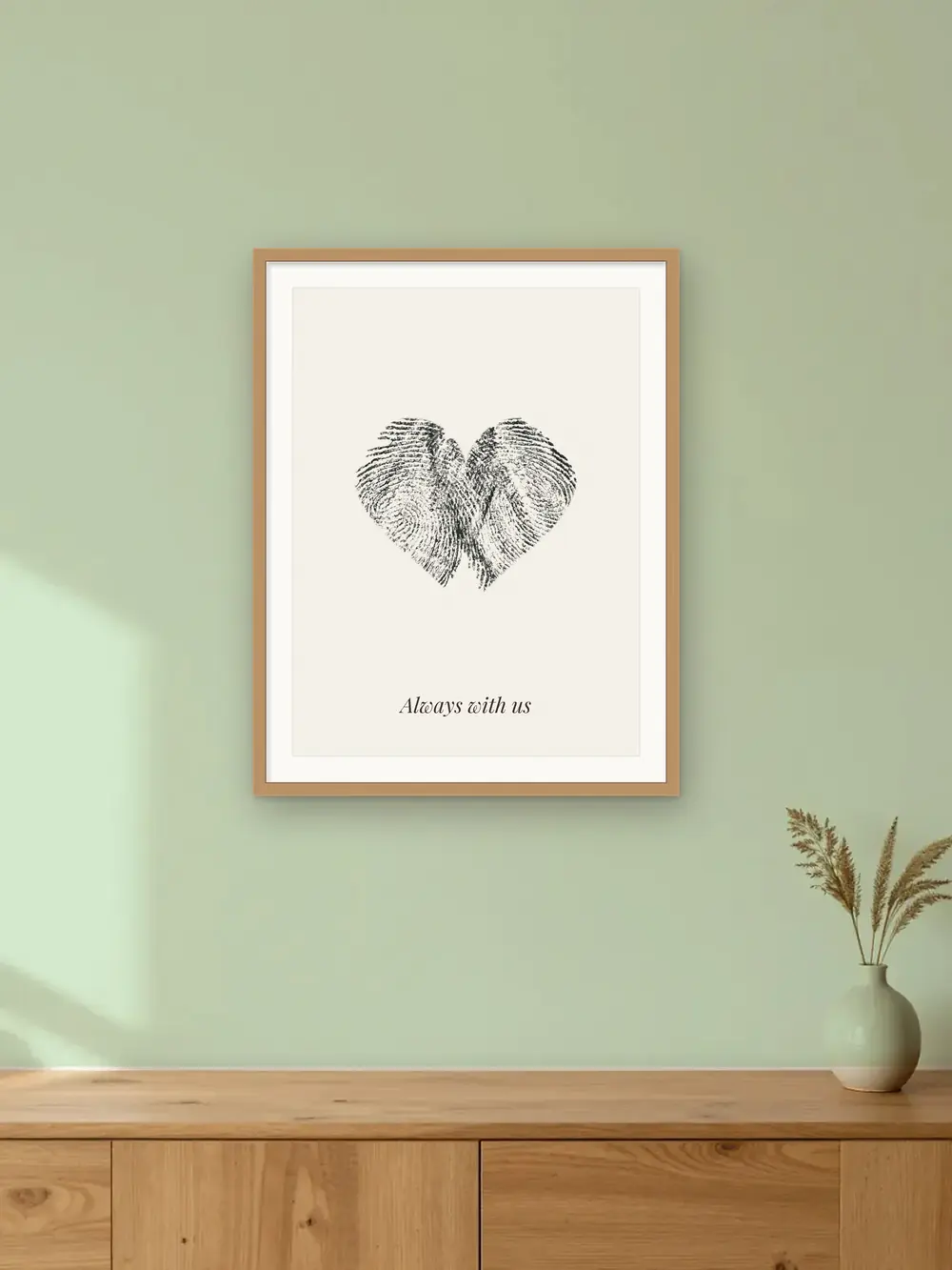 Memorial keepsake mockup: a fingerprint heart made from two real ink prints, in an oak frame on a calm sage wall labelled “Always with us”